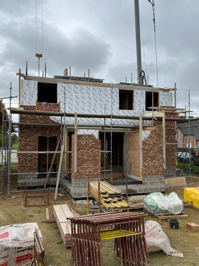 Woning in ruwbouw met steigers: gevelmetselwerk in rode baksteen op de begane grond en isolatieplaten op de verdieping
