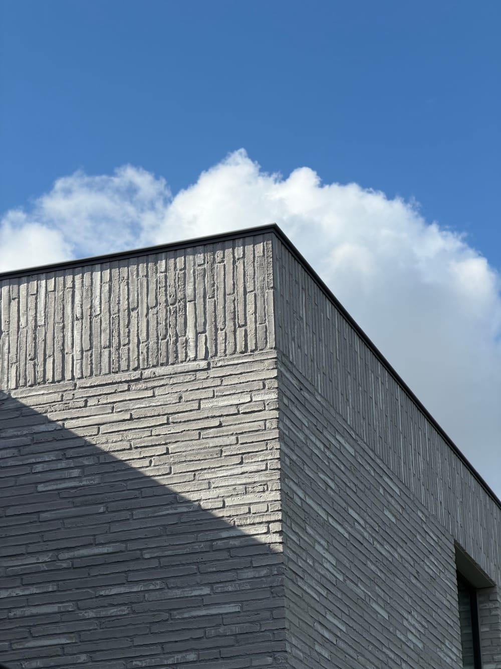 Hoekdetail van gevelmetselwerk in donkergrijze strengperssteen met een lichtere accentband onder de dakrand