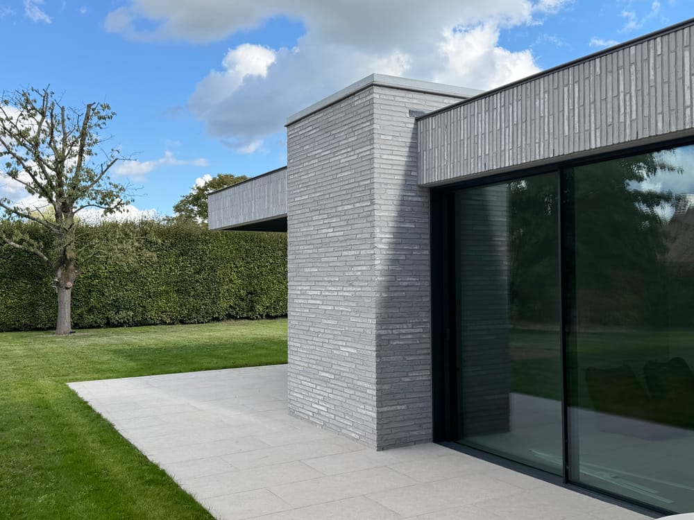 Moderne woning met plat dak in lichtgrijze gevelsteen, met schuiframen in zwart schrijnwerk en een terras aan de tuinzijde
